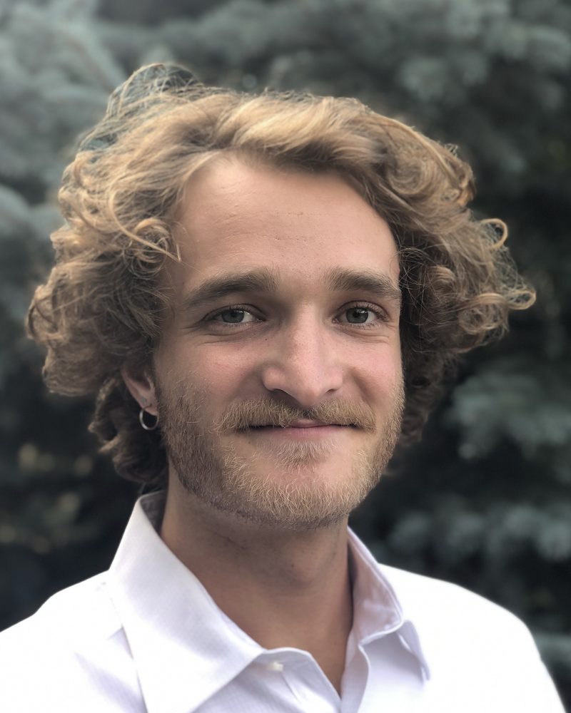 Jake Goulder, smiling, outdoors in front of evergreen trees